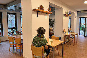 Eine nur von hinten zu sehende Frau sitzt an einem Tisch in einem großen, hellen Raum.