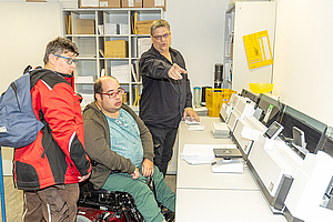 Daniel Schäfer, im Rollstuhl, und Sabrina Dudek schauen auf die große Frankiermaschine. Mitarbeiter Michael Geister erklärt.