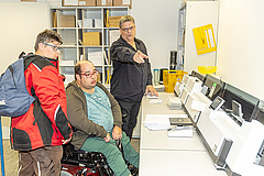 Daniel Schäfer, im Rollstuhl, und Sabrina Dudek schauen auf die große Frankiermaschine. Mitarbeiter Michael Geister erklärt.