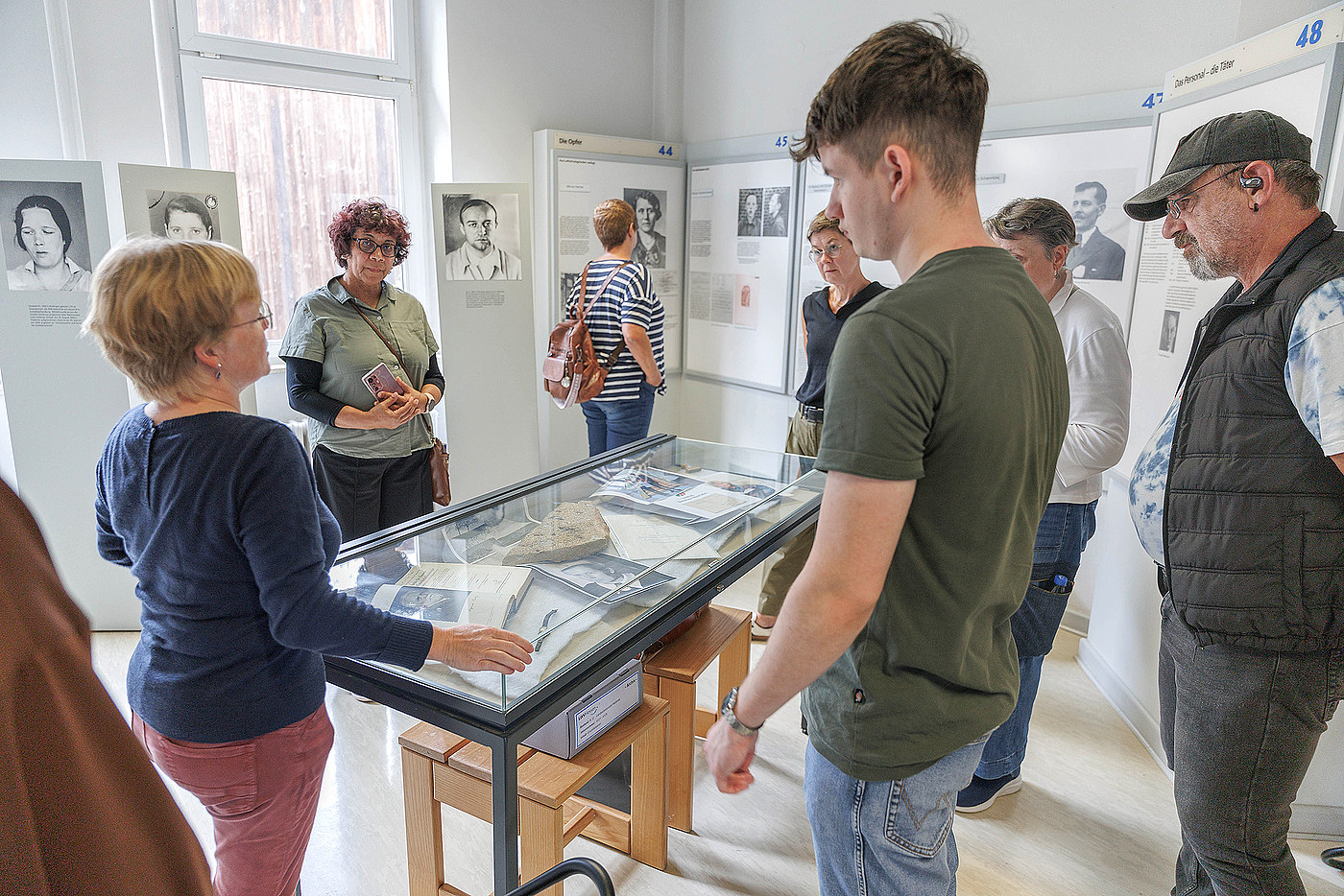 Eine Frau steht vor einer Ausstellungsvitrine mit Dokumenten unter Glas. Drunherum stehen sechs weitere Personen, Frauen und Männer von jung bis alt, und hören zu.