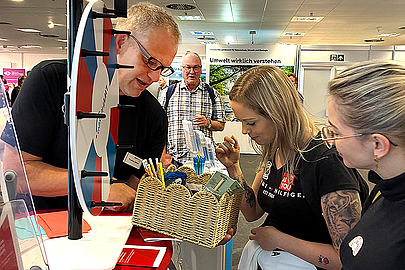 Kleine Gewinne winken: Das LWV Hessen Integrationsamt ist mit einem Glücksrad am Stand präsent. Ein Mann hält zwei jungen Frauen ein Körbchen mit Give-aways hin. Daneben ein Glücksrad.