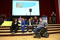 Thementalk (v.l.): LWV-Landesdirektorin Susanne Simmler, Staatsministerin Heike Hofmann, Moderator Bernd Bark und Landesbehindertenbeauftragter Andreas Winkel. Auf der Bühne die inklusive Band Saitenwind der bdks, rechts Gebärdensprachdolmetscherin Antj Die vier Teilnehmen des Thementalks sitzen auf der Treppe zur Bühne im Ständesaal
